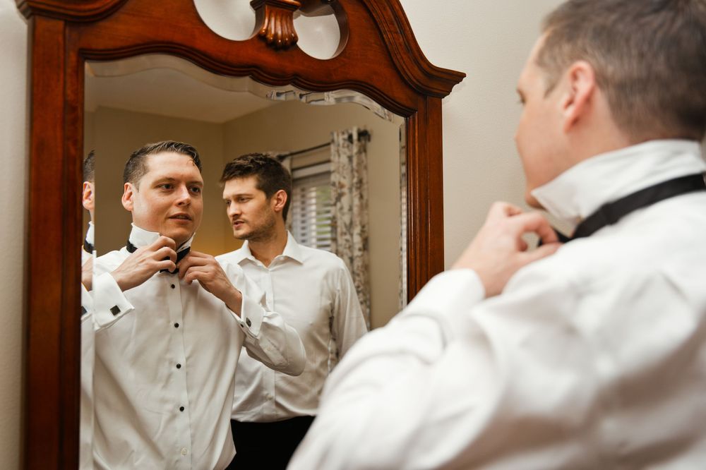 Two men in white dress shirts preparing for a wedding, adjusting ties while looking in an ornate wooden mirror.