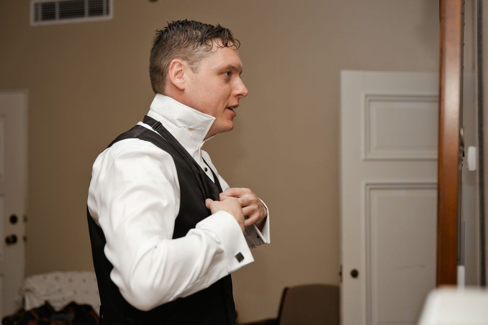 Groom adjusting his bow tie while wearing a white shirt and black vest during wedding preparations.