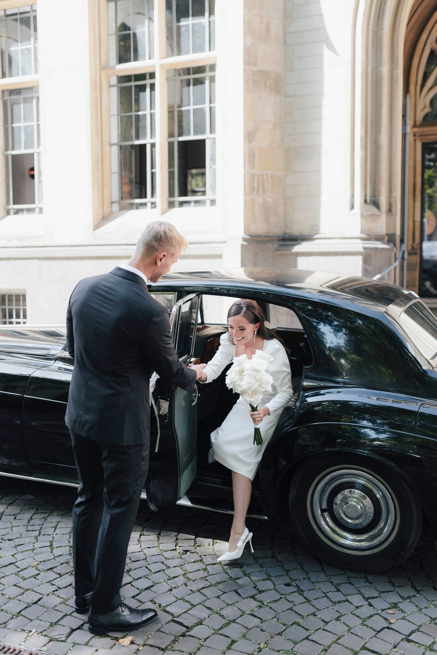 Groom in black suit helps bride in white dress exit a classic black luxury car outside an elegant building.