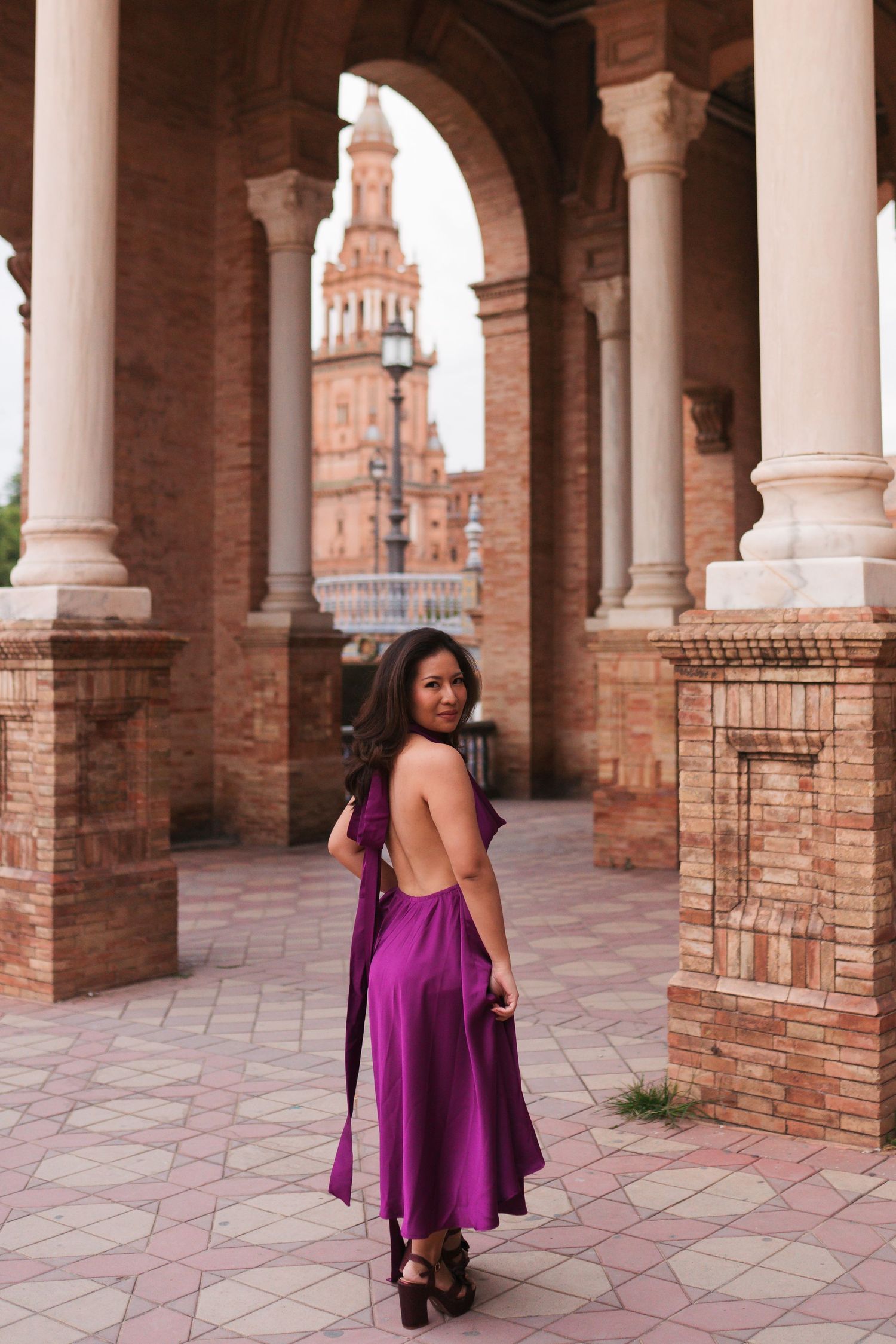 Woman in elegant backless purple dress posing under arched colonnade at Plaza de España, Seville, Spain.