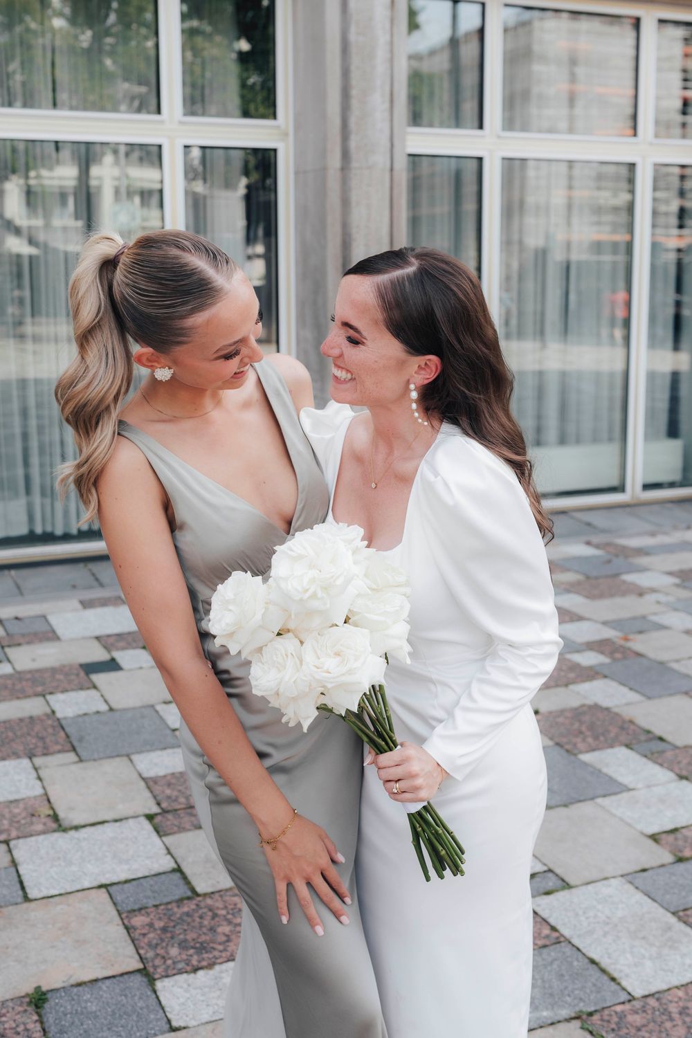 Bride in white satin dress holding white peony bouquet smiling with bridesmaid in silver gown outdoors.