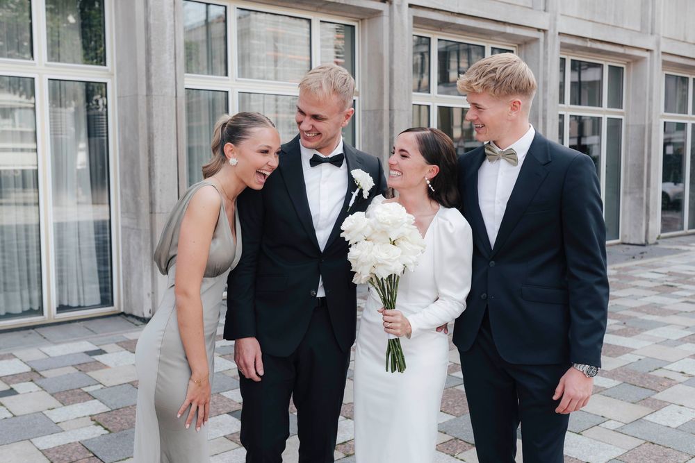 Wedding party of four posing outdoors, two men in tuxedos and two women, one in a white bridal gown holding flowers.