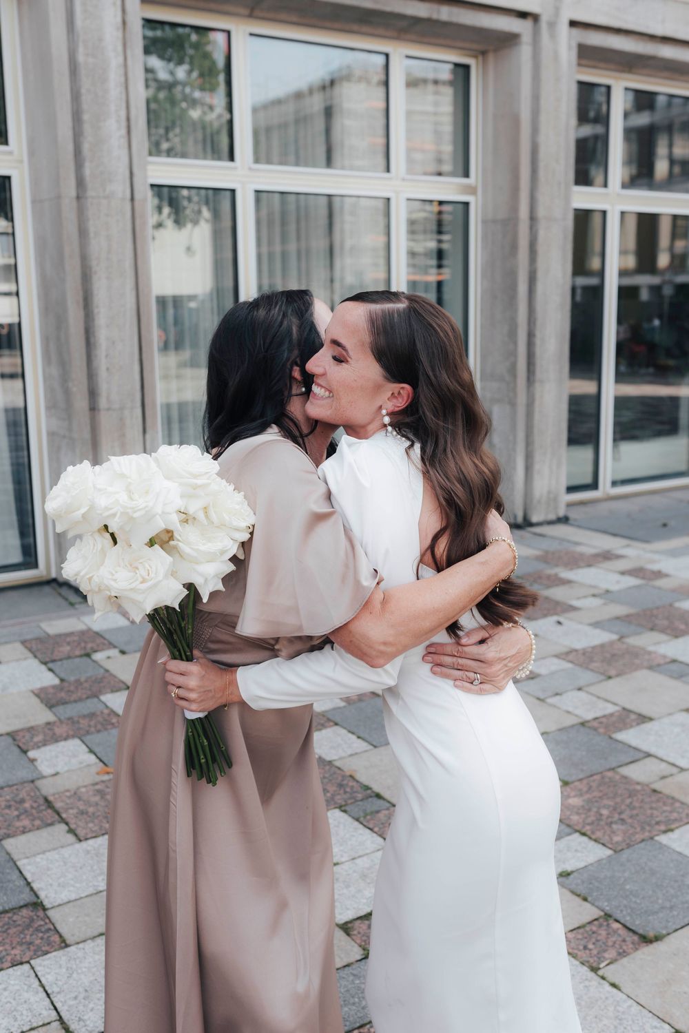 Two women in elegant dresses embracing outdoors, one in white bridal gown holding white roses bouquet.