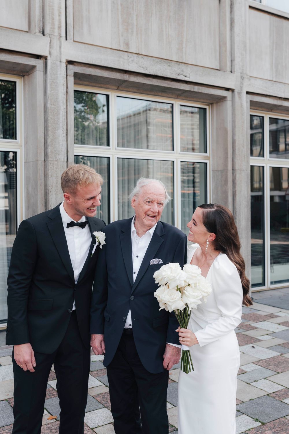 A bride and groom in formal wedding attire smile with an elderly man outside a modern building.