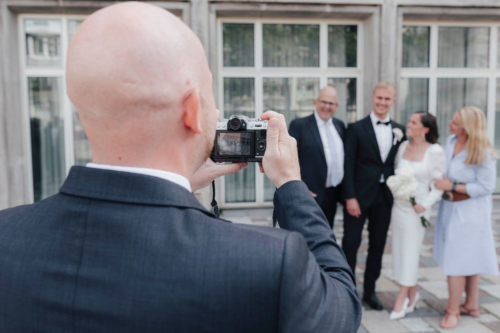 Bald photographer capturing wedding party portrait outdoors, bride in white holding bouquet with guests smiling.