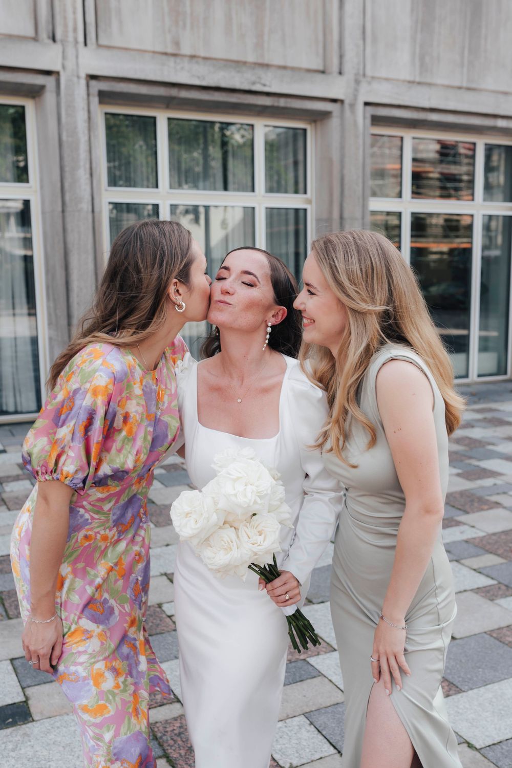 Bride in white dress holds bouquet while two friends in colorful and silver dresses kiss her cheeks outdoors.