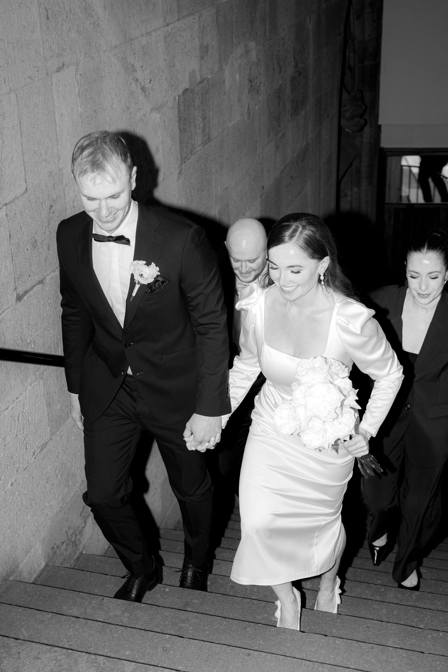 Black and white photo of a bride and groom holding hands walking up stone steps at their wedding.