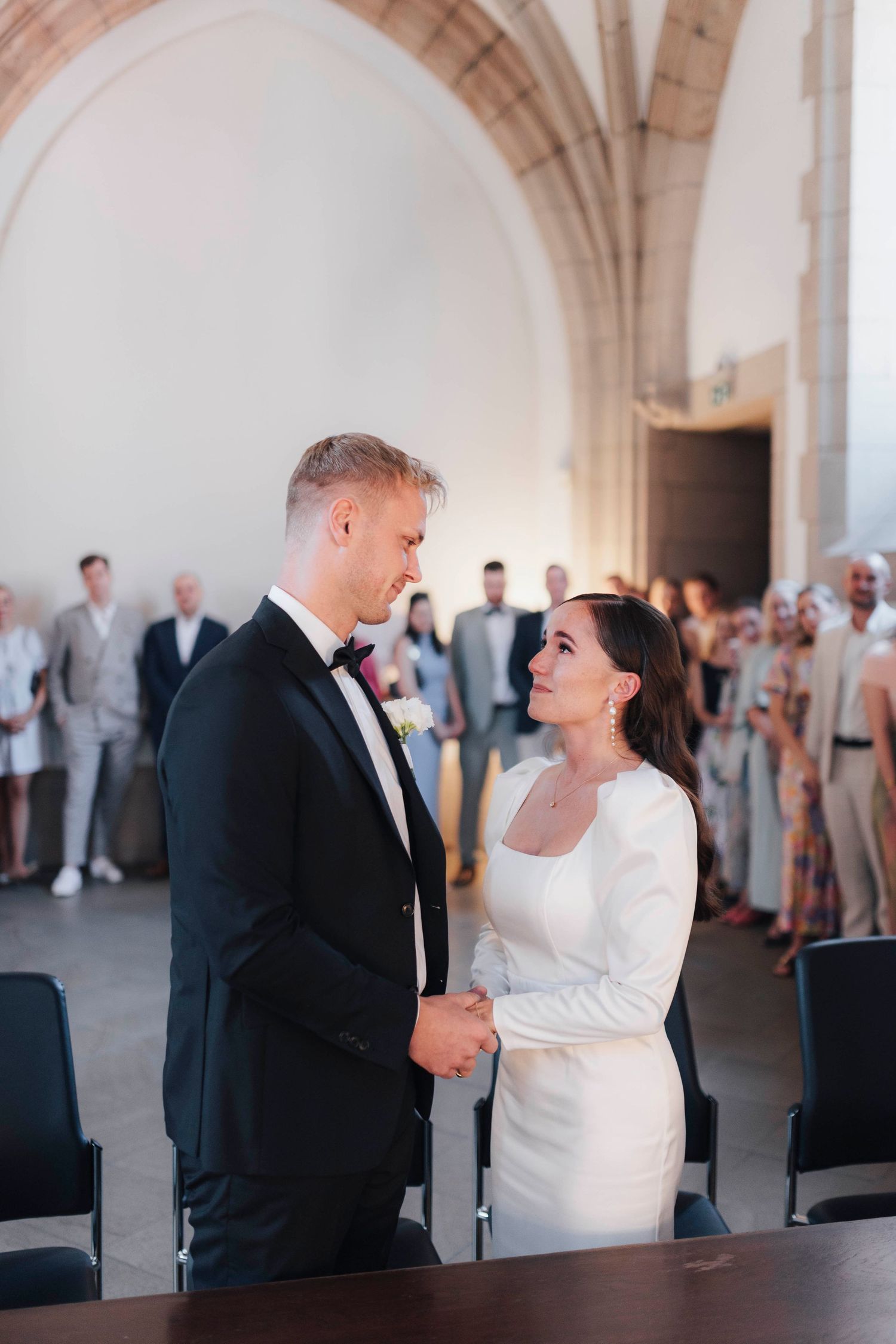 A couple exchanges vows at an elegant wedding ceremony in a grand arched hall with guests watching.