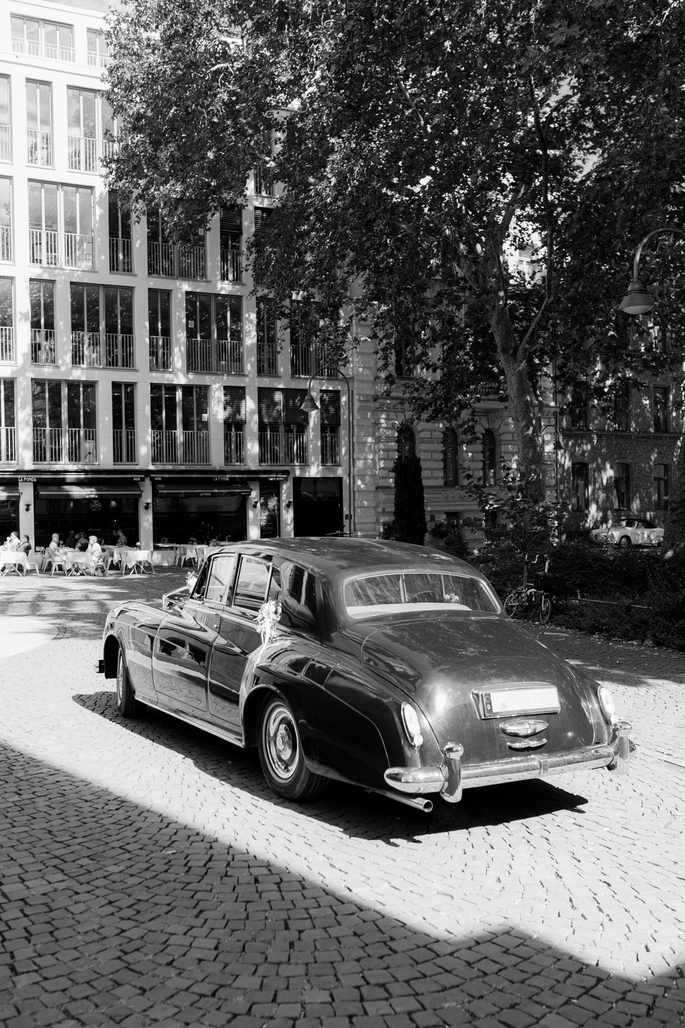 Vintage classic car parked on cobblestone street in black and white, with trees and modern building in background.