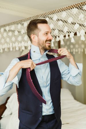 A person in formal attire adjusting a burgundy tie while getting ready.