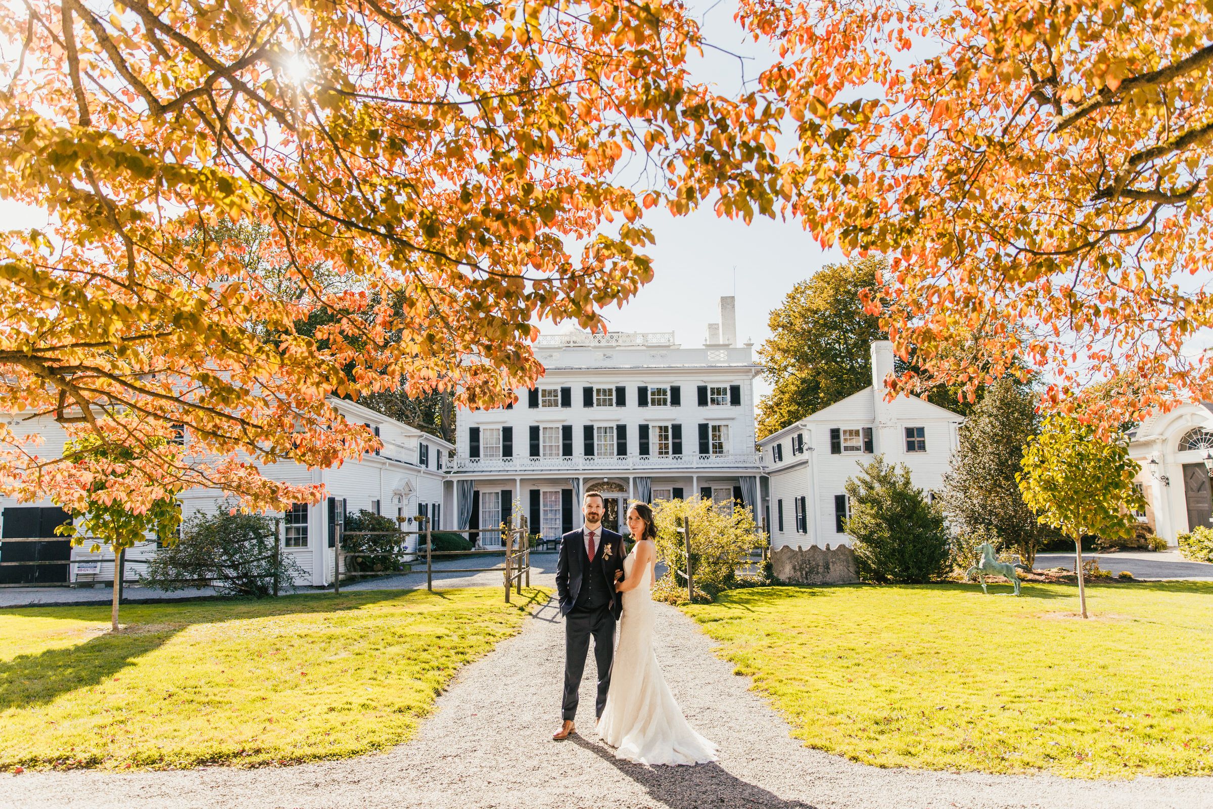 A couple walking on a path towards a white colonial mansion surrounded by autumn trees.