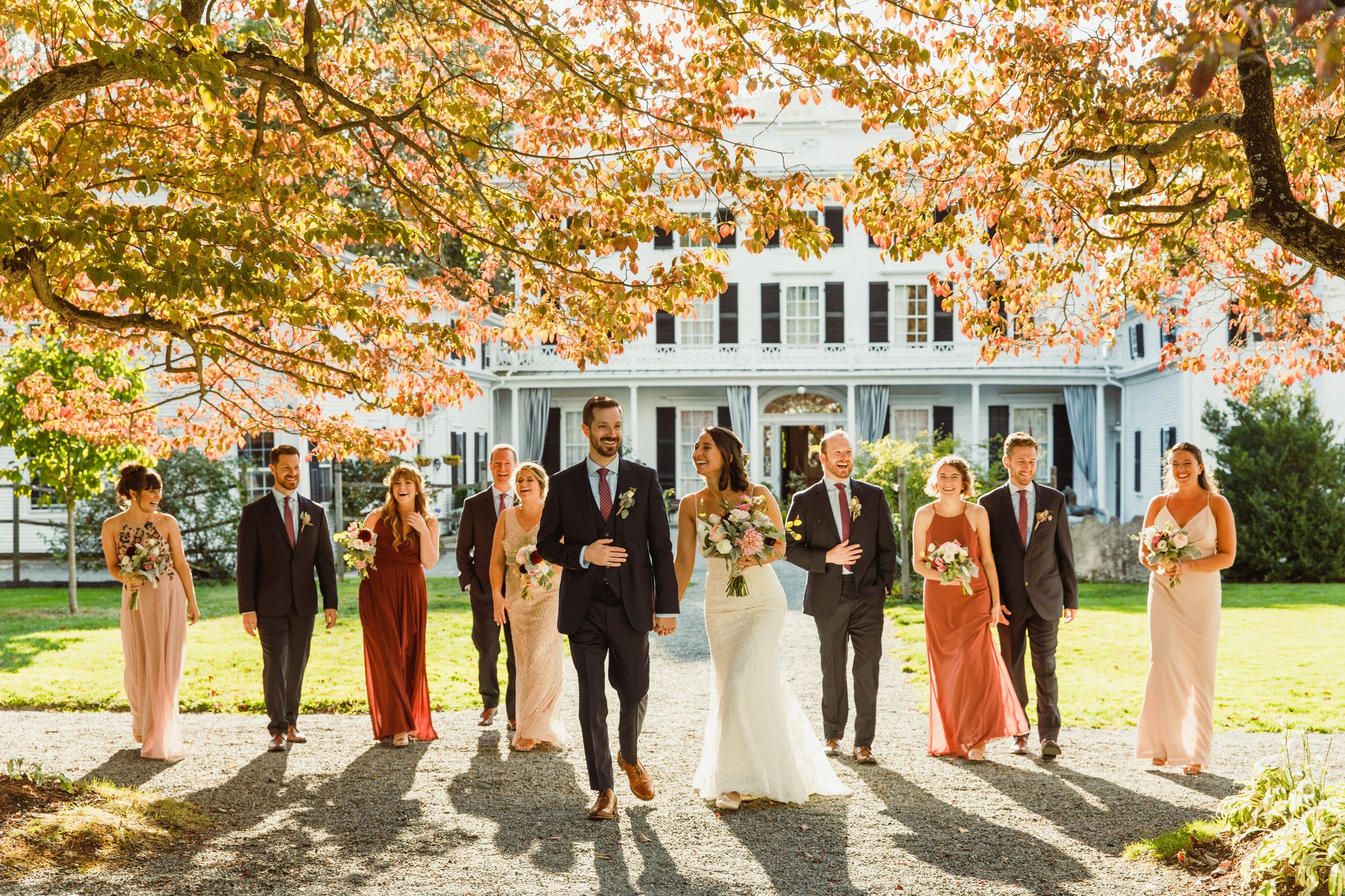 Wedding party posed in front of a grand white mansion during fall season.