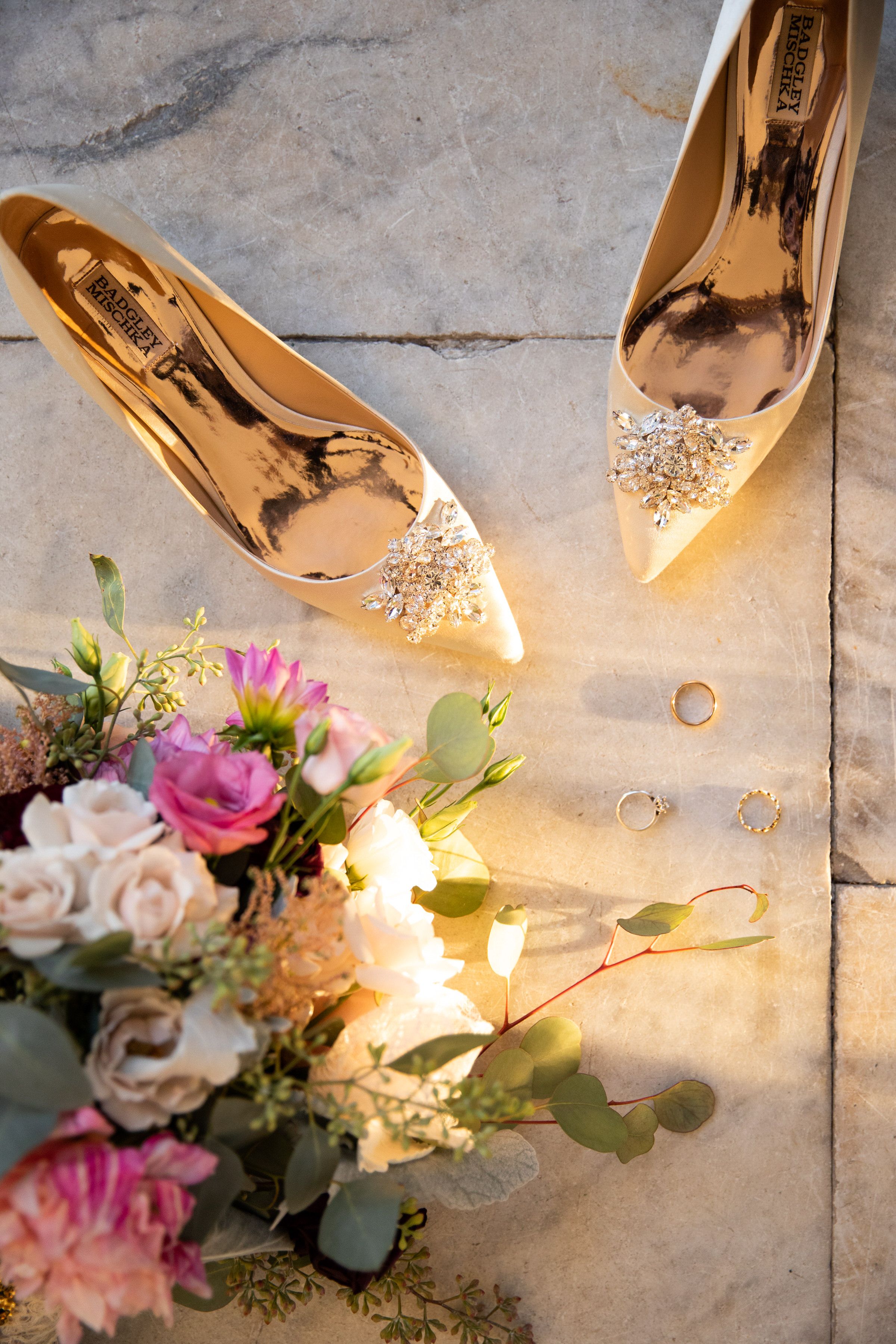 Elegant gold bridal shoes with crystal embellishments next to a pink and white floral bouquet on stone flooring.