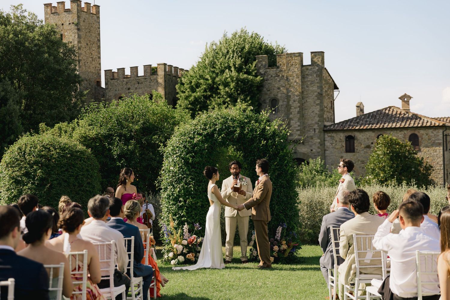 Outdoor wedding ceremony on a lawn with guests seated in white chairs before a floral arch, with a castle backdrop.