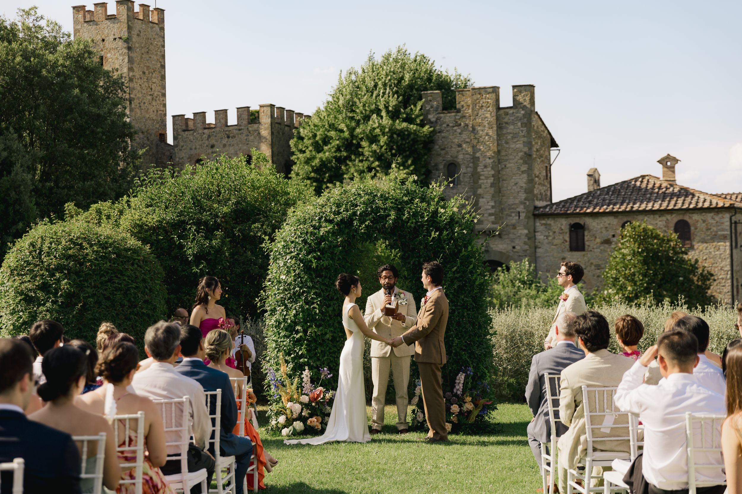 Outdoor wedding ceremony on a lawn with guests seated in white chairs before a floral arch, with a castle backdrop.