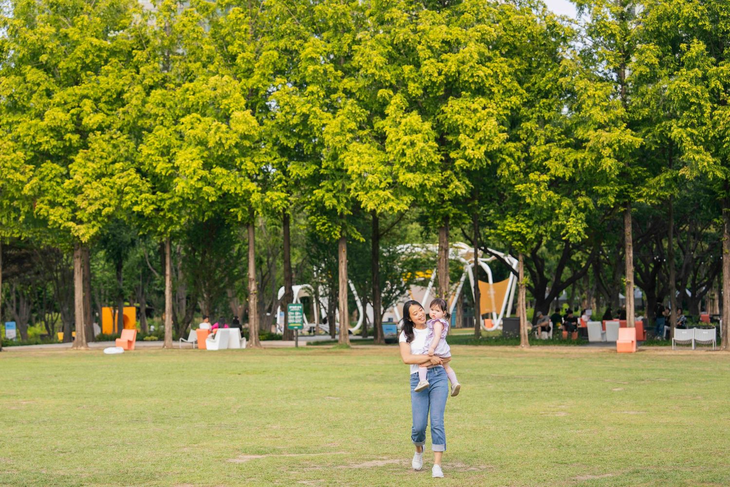 A woman holds a child in a lush green park surrounded by tall trees on a sunny day.