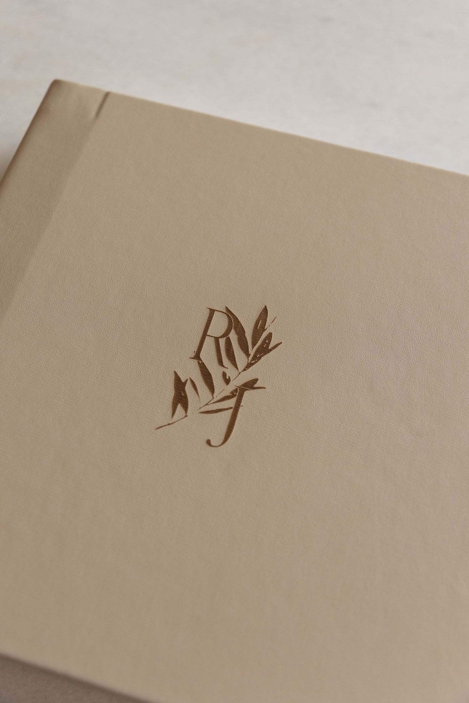 Elegant beige linen book with gold embossed monogram and botanical leaf design, perfect for wedding guest books.