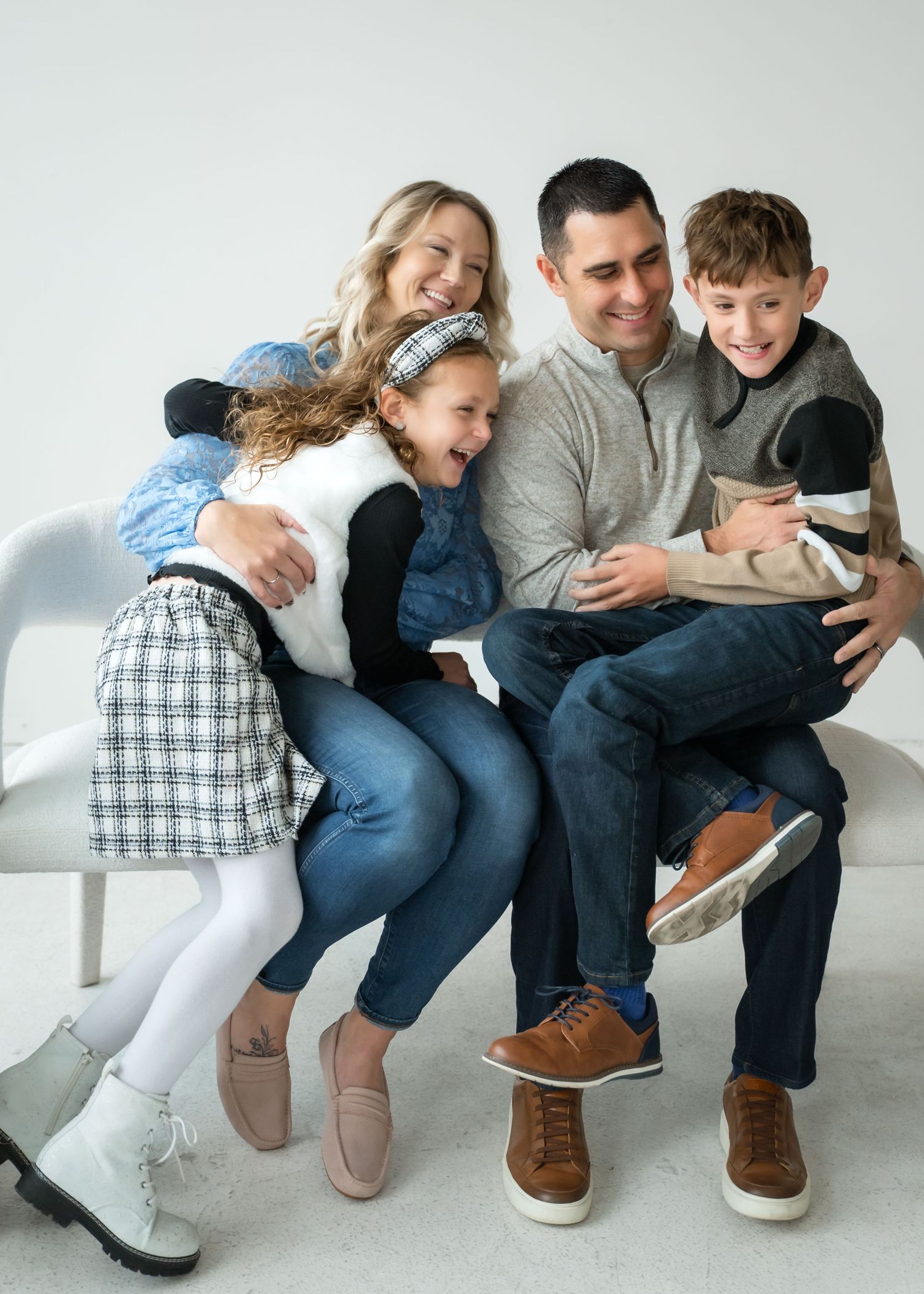 Happy family of four laughing together on a white bench in a bright studio portrait setting.