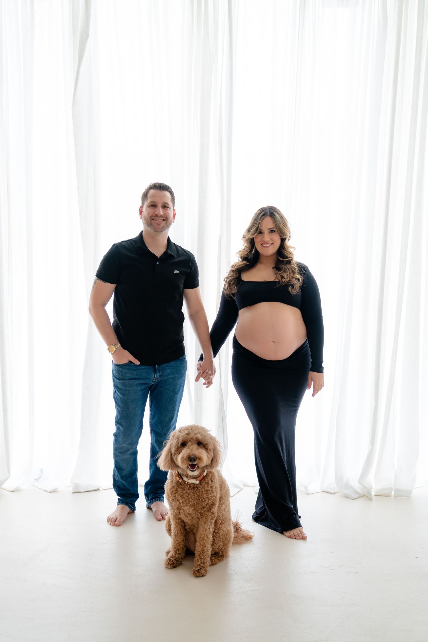 Couple in black outfits holding hands with golden doodle dog during maternity photo session against white curtains.
