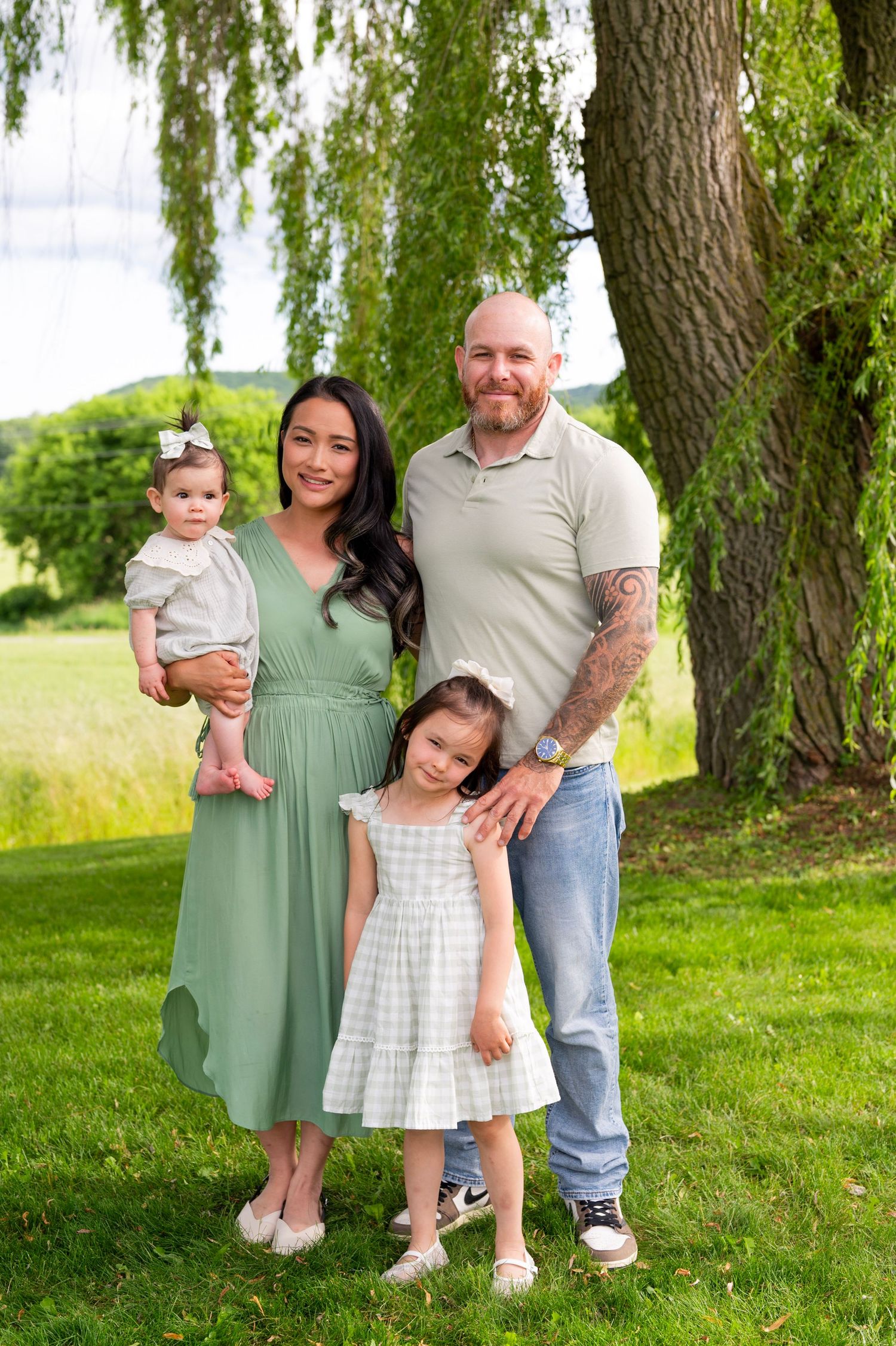 Two women and two young girls smile together on green grass under a willow tree on a sunny day.