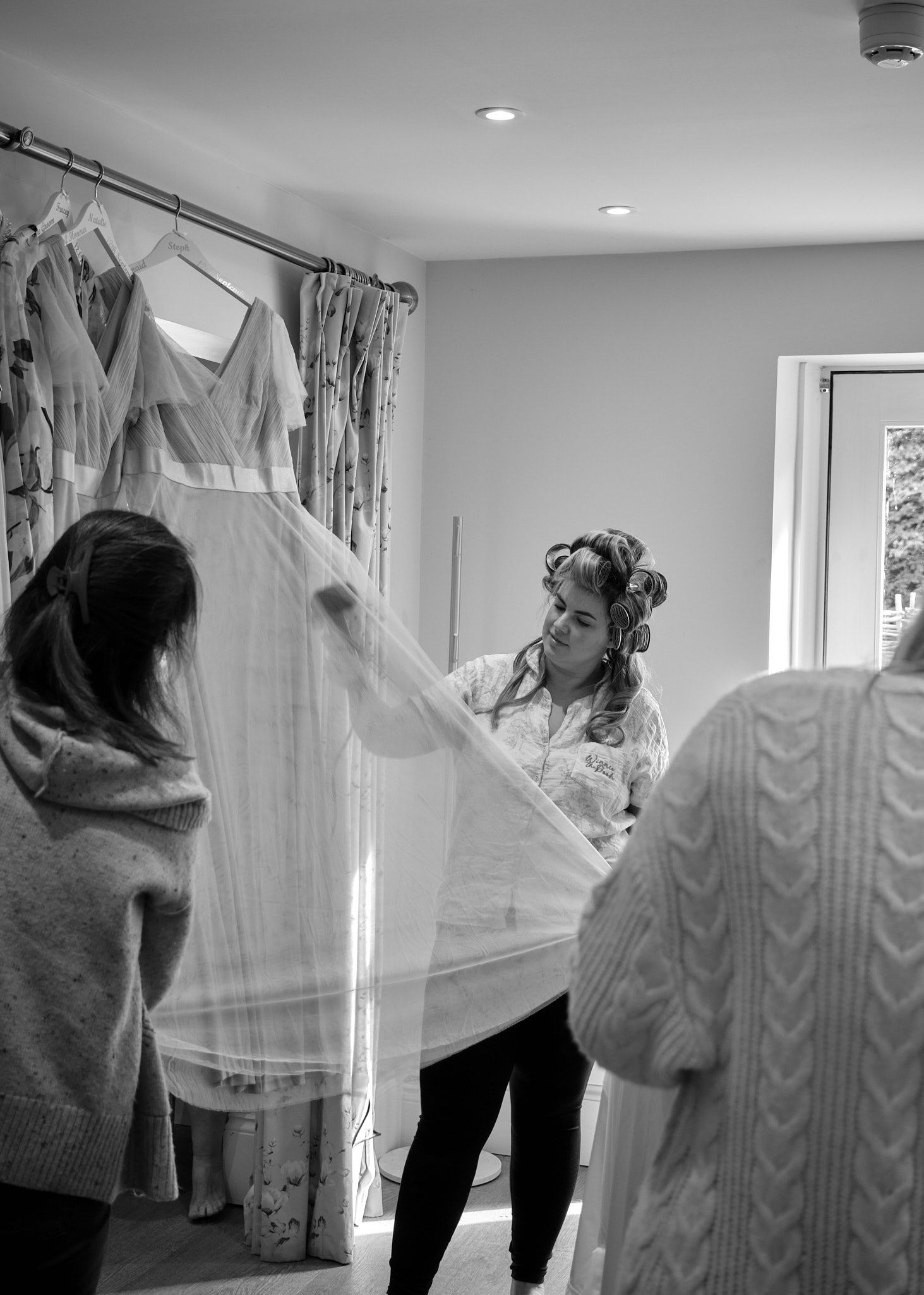 Black and white photo of women handling a white wedding dress in a bridal fitting room with gowns hanging in background.