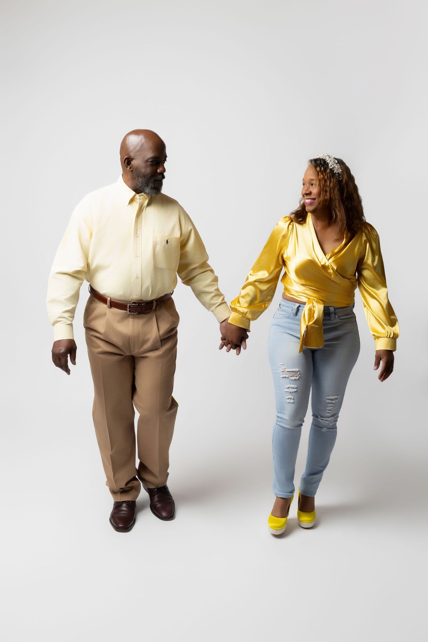 A smiling Black couple holds hands and walks together, wearing coordinating yellow outfits with tan pants and light blue jeans.