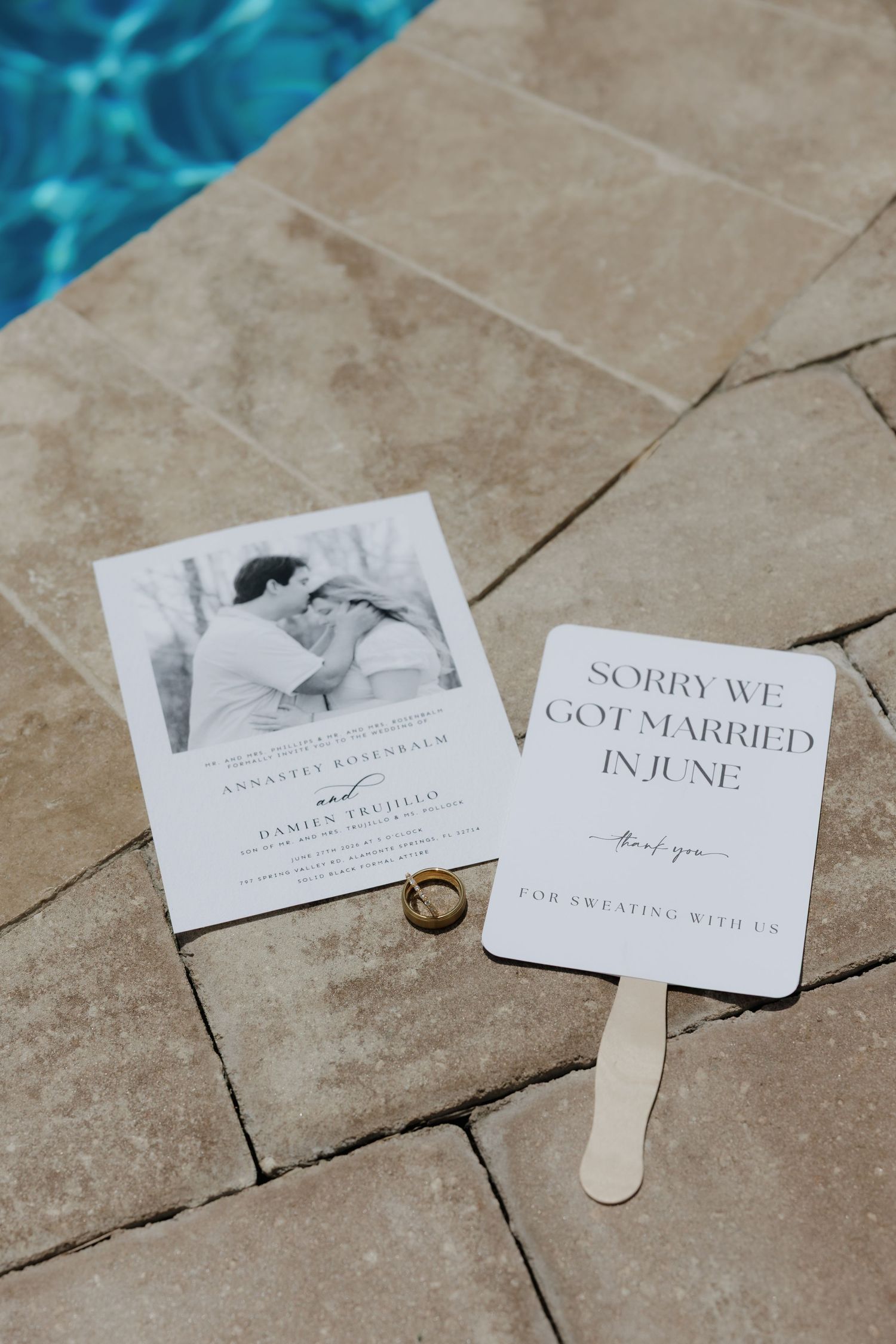Wedding invitation, fan program, and rings displayed on stone tiles beside a sparkling blue pool.