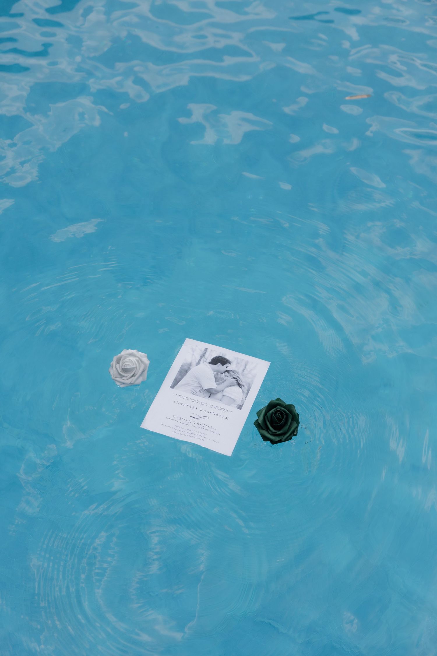 Wedding invitation card floating in a blue swimming pool with two decorative roses beside it.