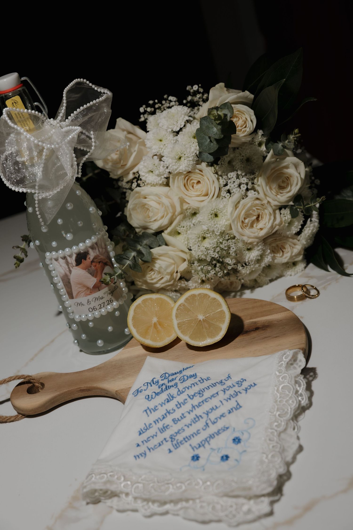 Wedding flat lay with white rose bouquet, gold rings, lemon halves, personalized bottle, and embroidered handkerchief on wooden board.