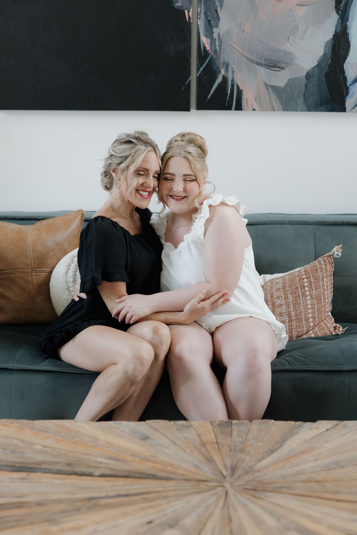 Bride in white romper and woman in black dress share joyful embrace on gray couch.