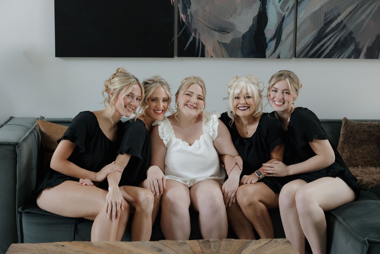 Bride in white with four bridesmaids in black dresses sitting together on a couch, smiling and laughing.