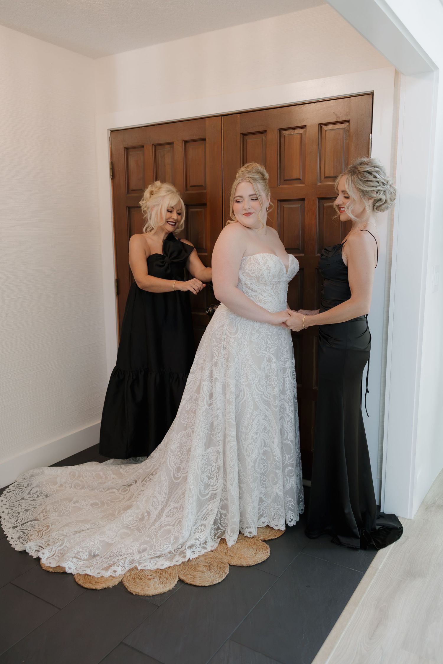 Bride in lace wedding gown assisted by two bridesmaids in black dresses near wooden doors.
