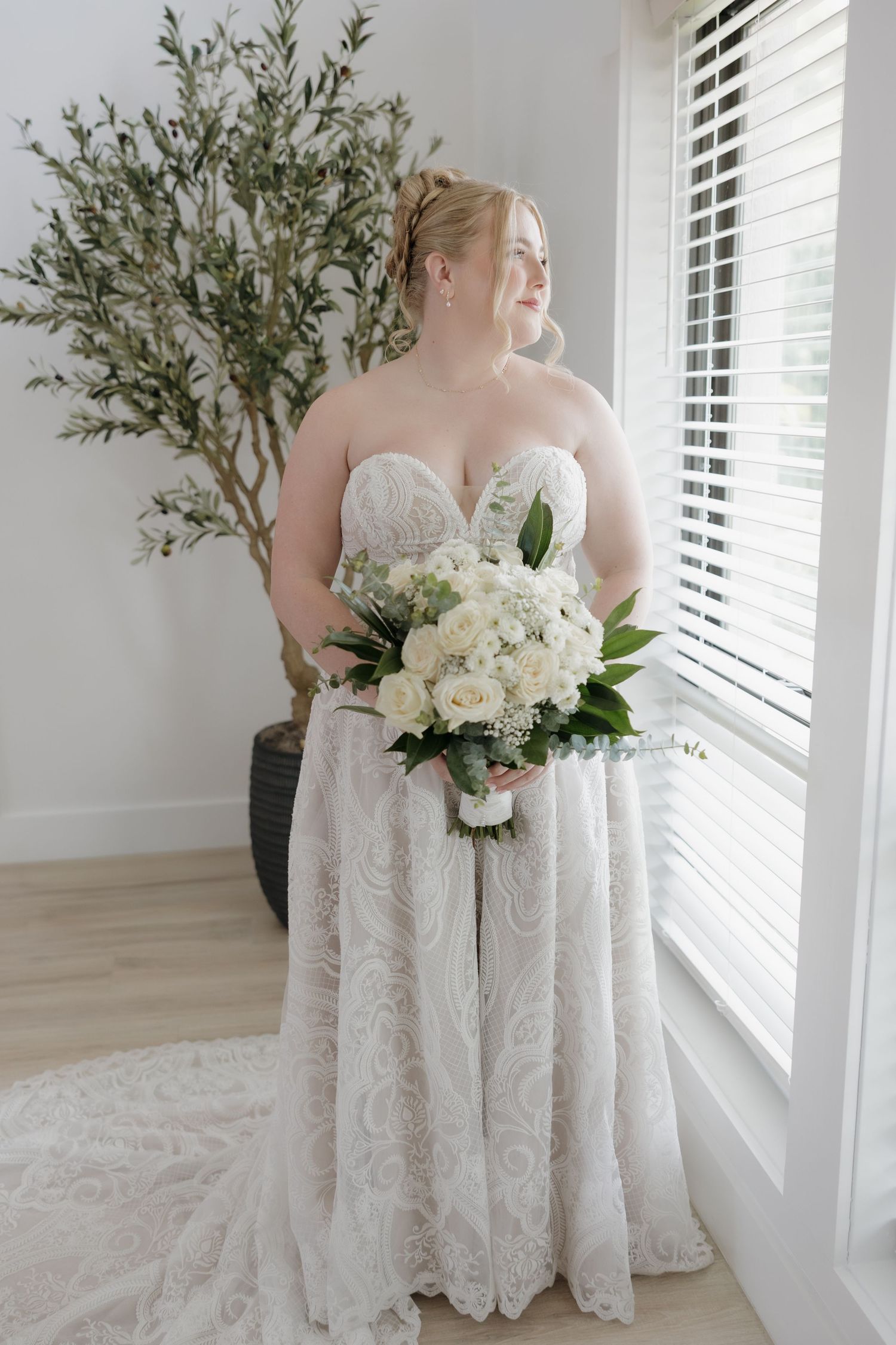 Bride in strapless lace wedding dress holding white floral bouquet near bright window with shutters.