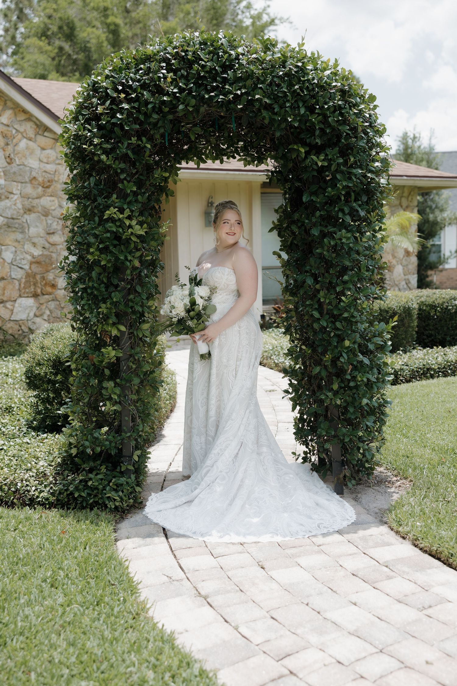 Bride in lace gown holds white bouquet standing under a lush green garden arch on a brick pathway.