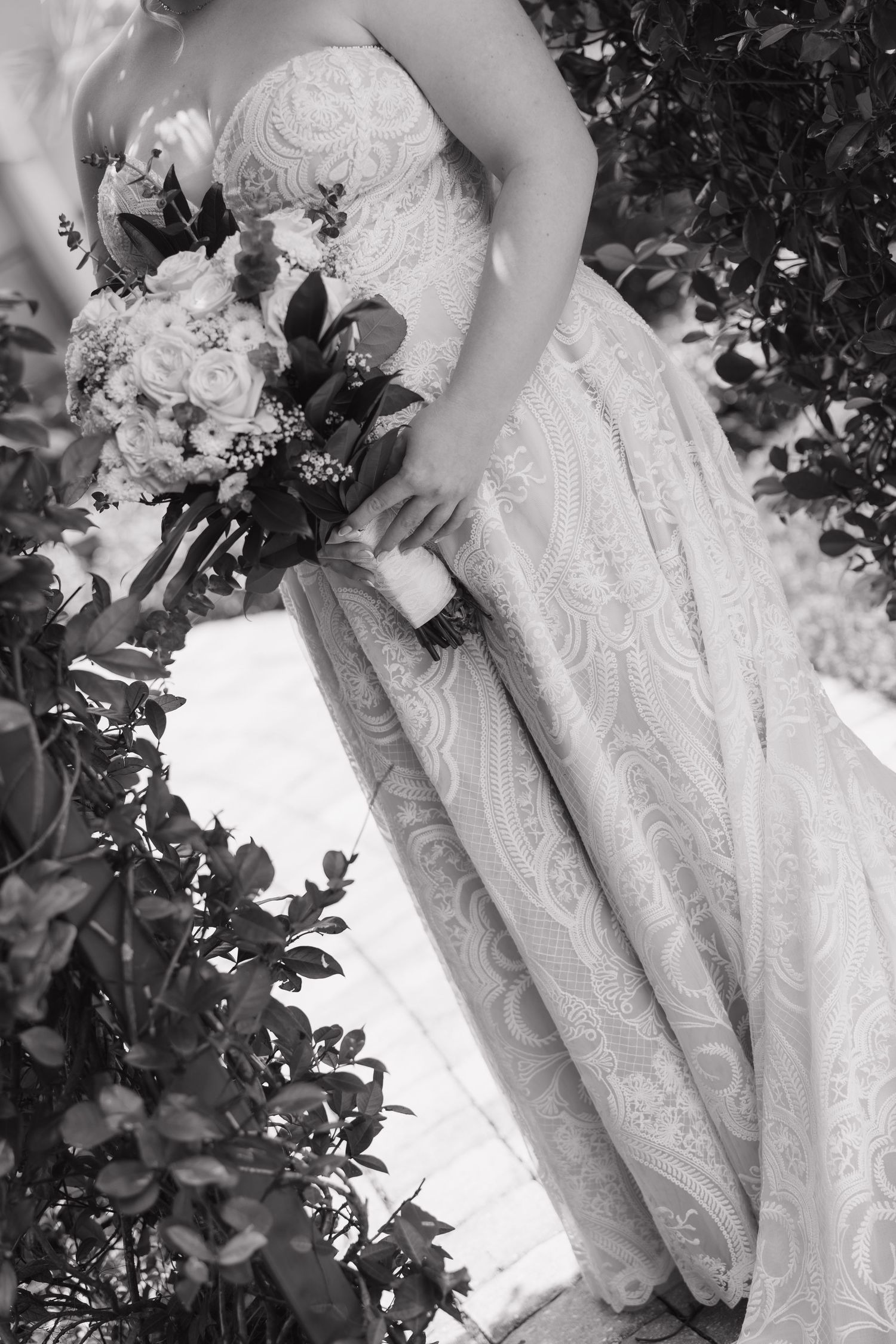 Black and white close-up of bride in lace wedding gown holding a beautiful floral bouquet outdoors.