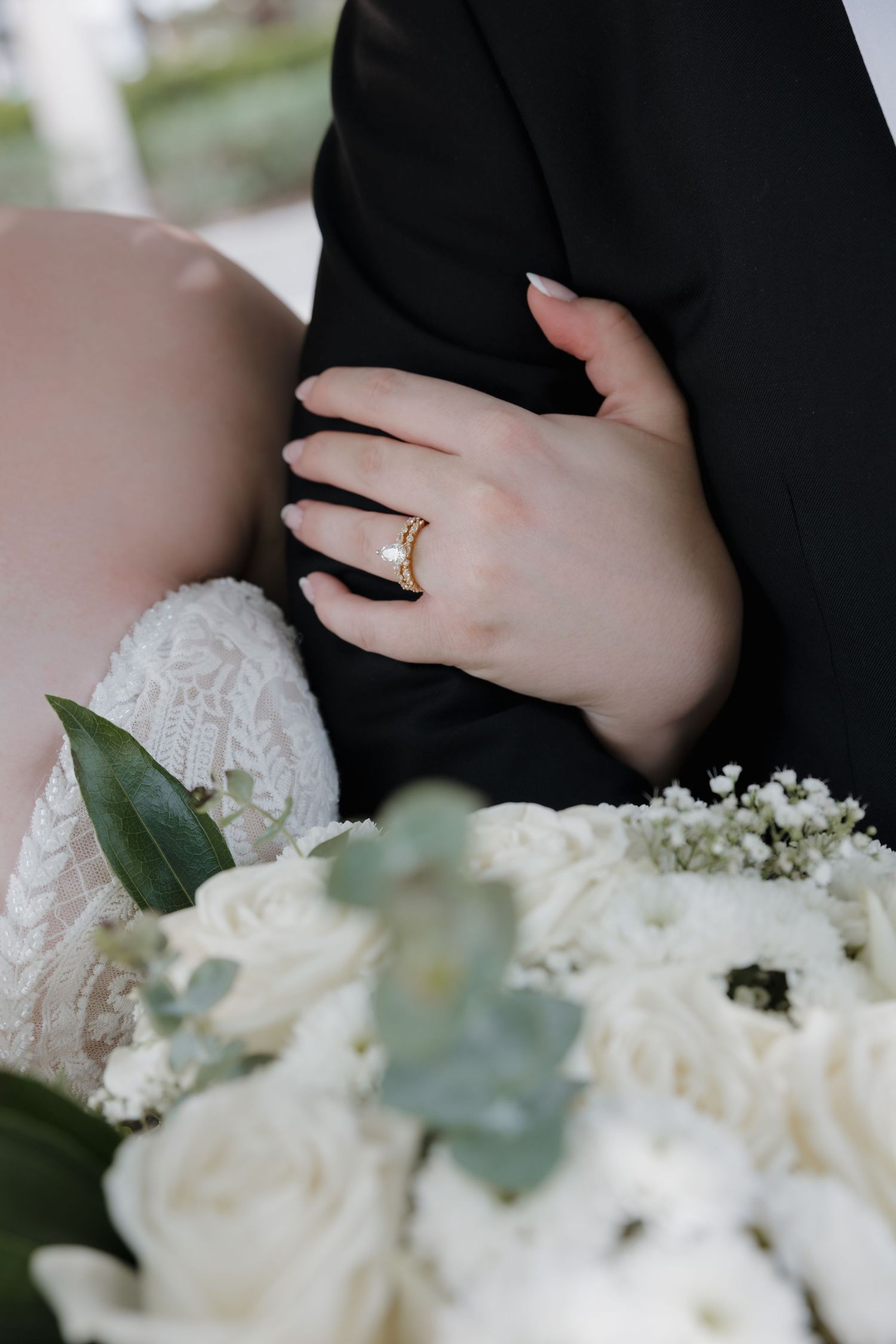Bride wearing a gold pearl ring holds groom's arm in black suit, with white rose wedding bouquet in foreground.