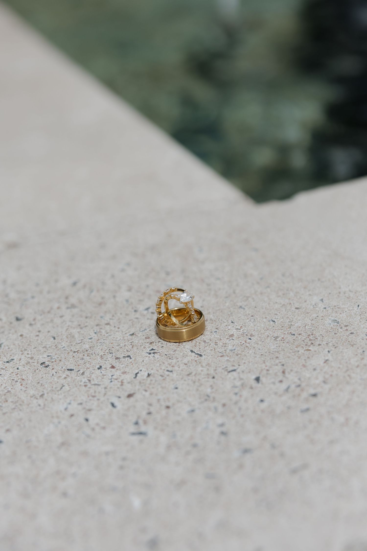 Two gold wedding rings, including a diamond engagement ring, rest on a concrete surface near a pool.