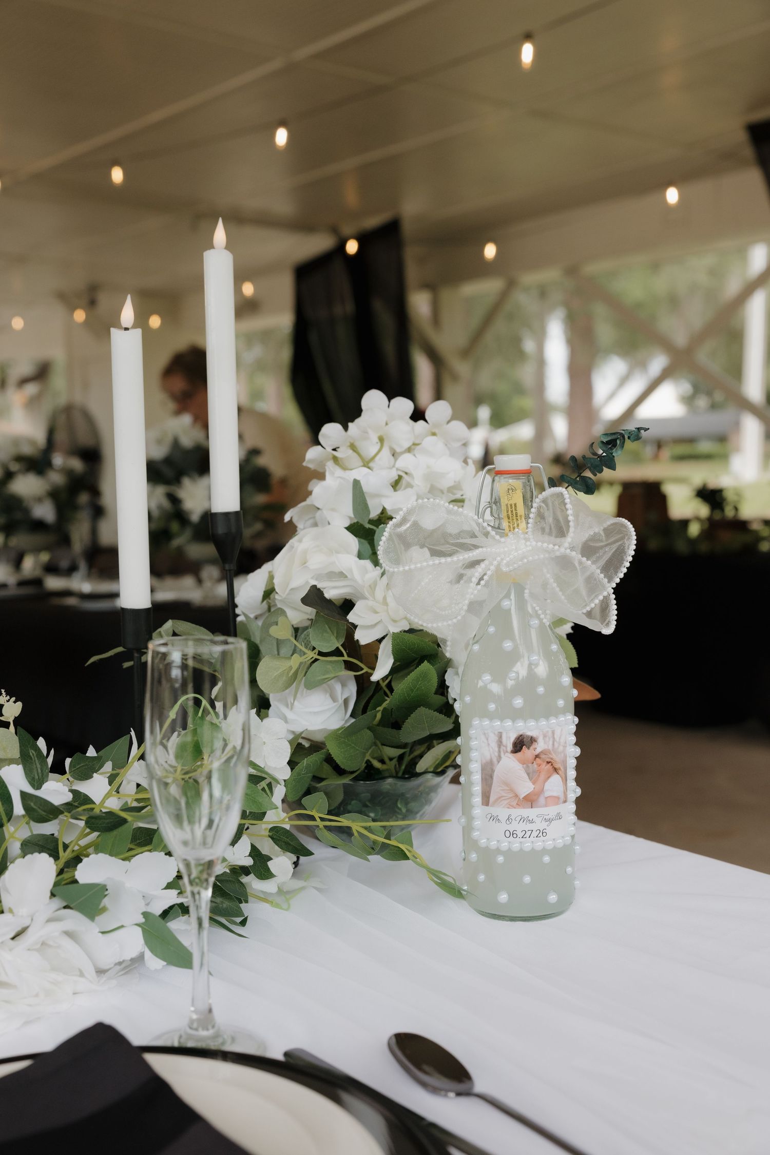 Elegant wedding reception table with white candles, floral centerpiece, and personalized bottle favor.