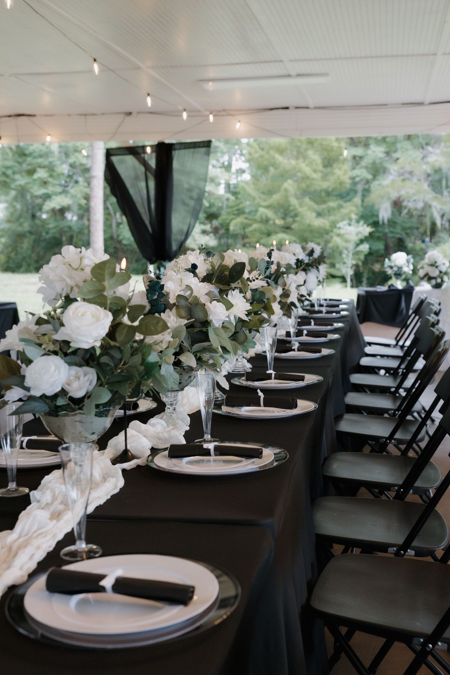 Elegant wedding reception table with black linens, white floral centerpieces, and string lights under a tent.