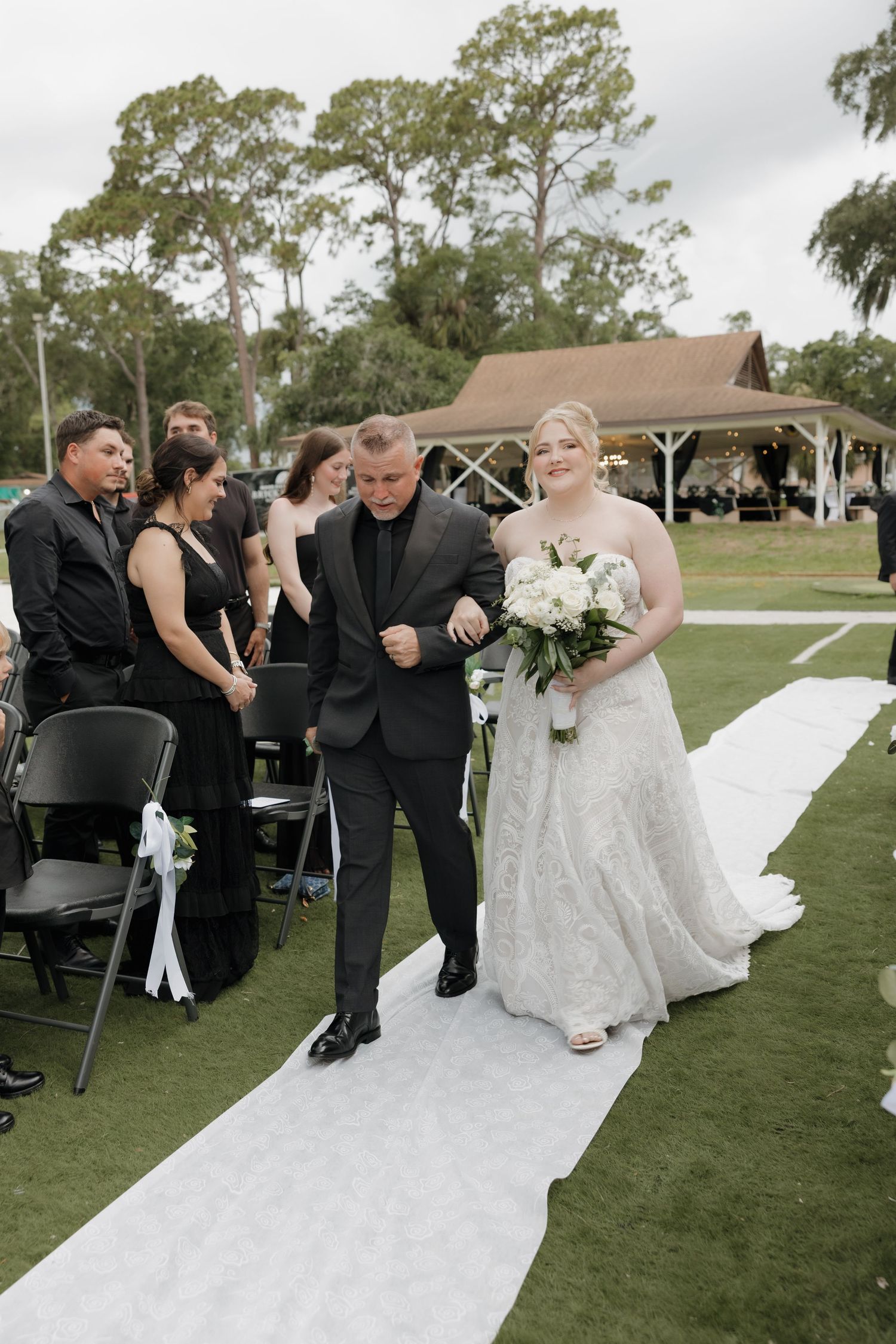 Bride in lace gown walks down white aisle with escort at outdoor wedding ceremony.