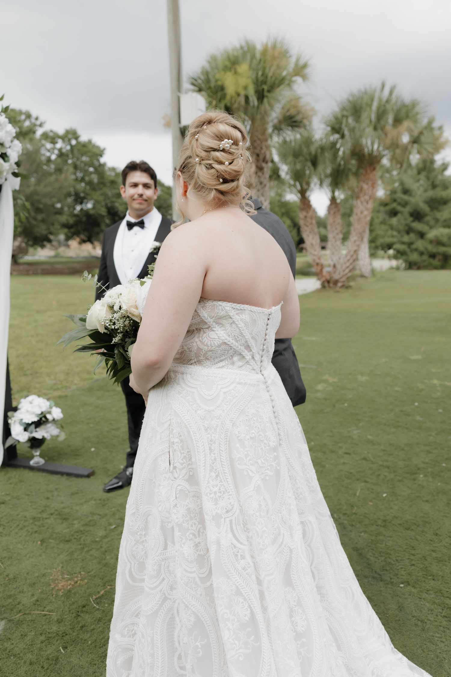 Bride in lace gown approaches smiling groom at outdoor wedding ceremony on green lawn.