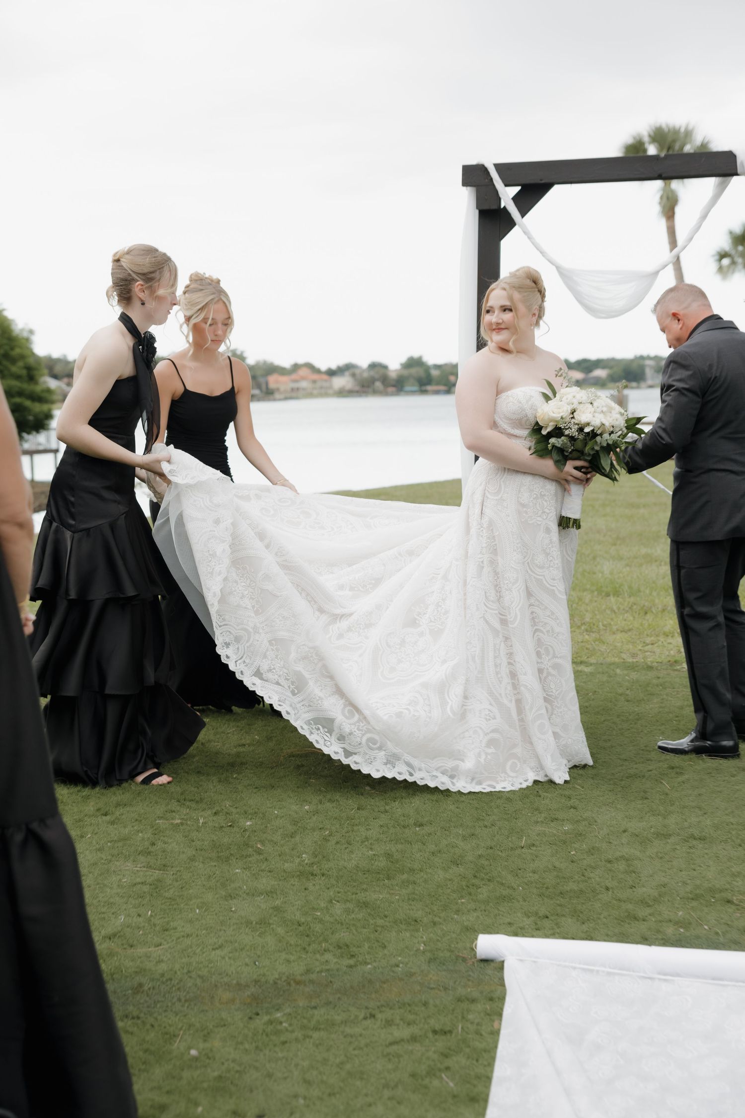 Bridesmaids in black dresses help bride with white lace wedding gown outdoors near waterfront ceremony arch.