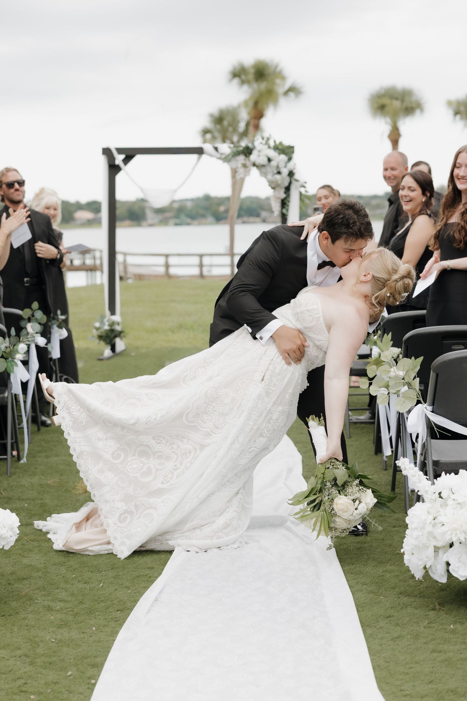 Groom dips bride for a kiss at outdoor waterfront wedding ceremony with guests cheering alongside.