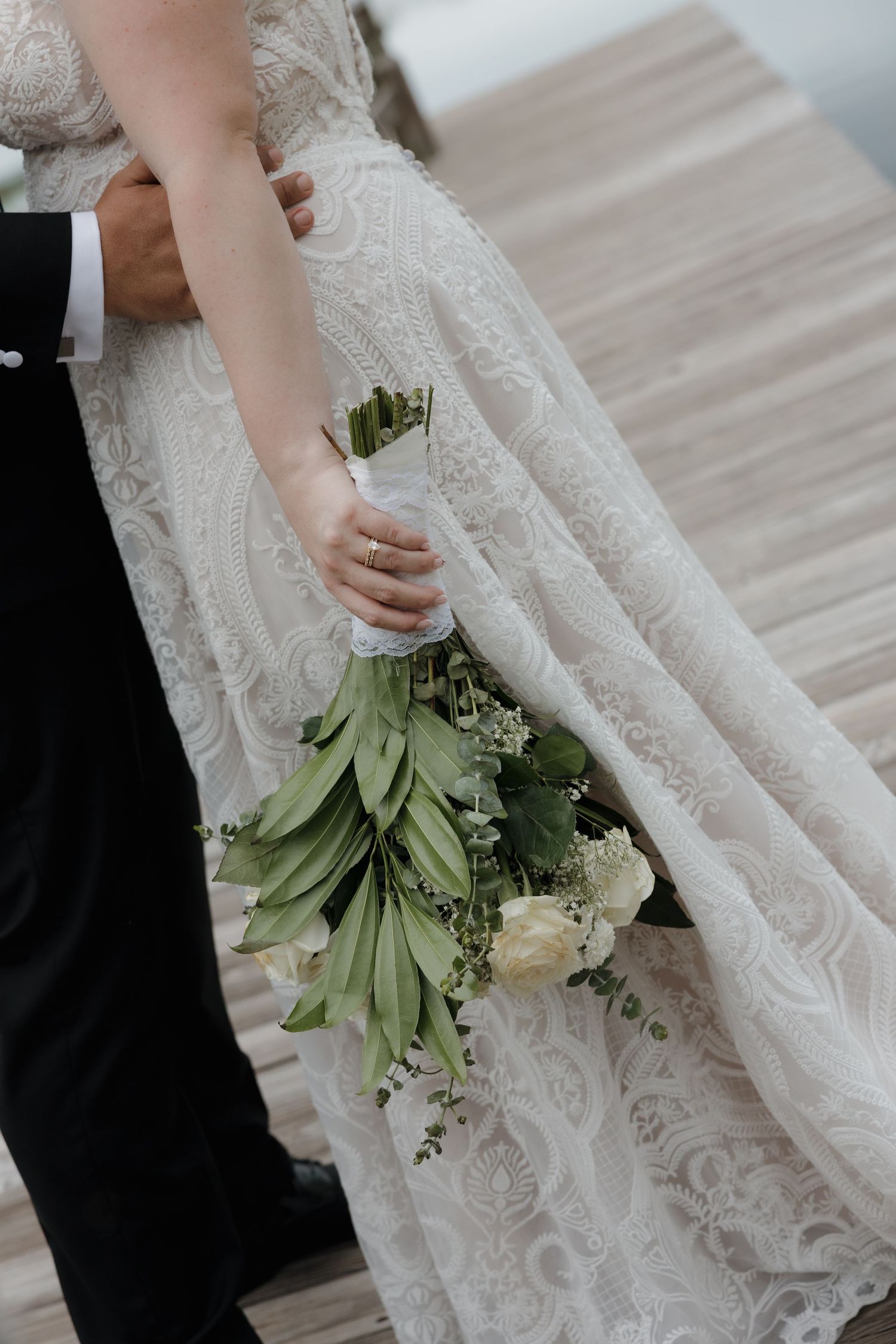Close-up of bride in lace gown holding green and white bouquet beside groom in tuxedo.