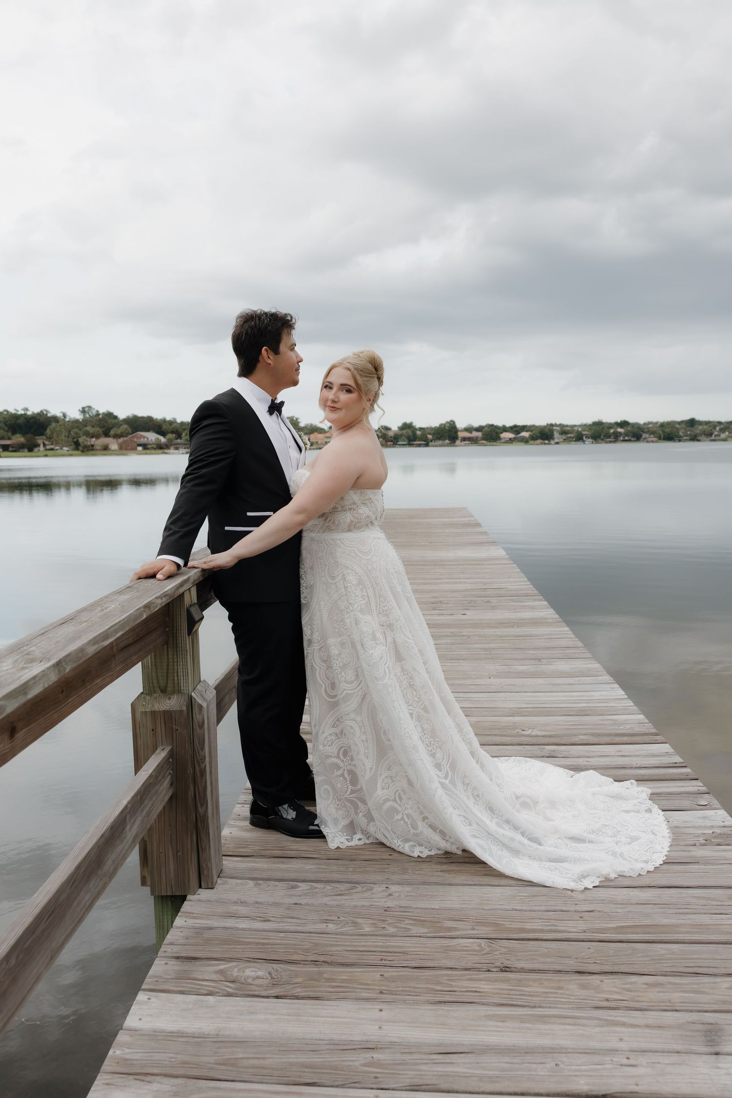 Bride and groom standing on wooden dock overlooking calm lake under cloudy sky.