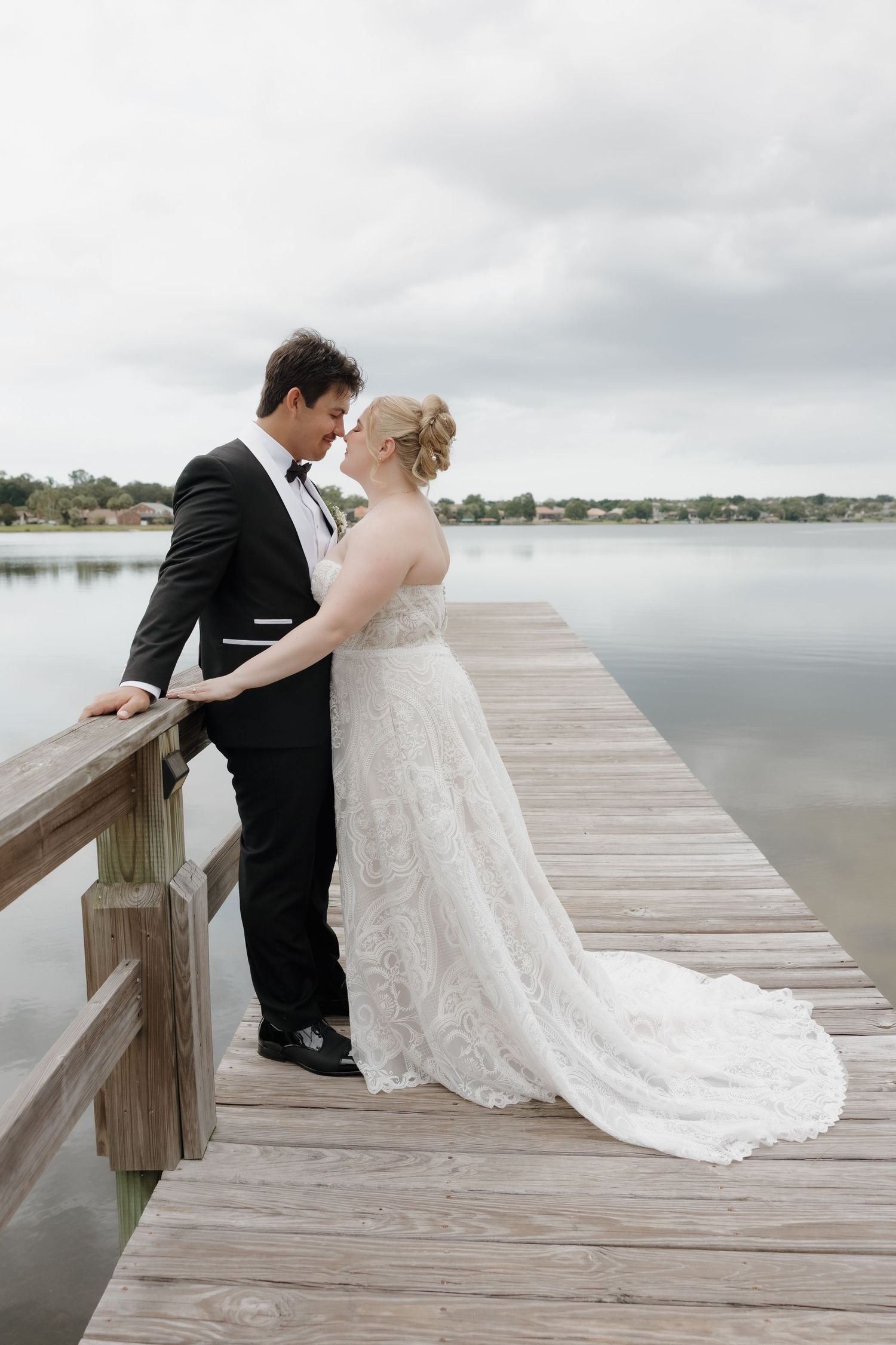 Bride and groom share a romantic moment on a wooden dock overlooking a calm lake on their wedding day.