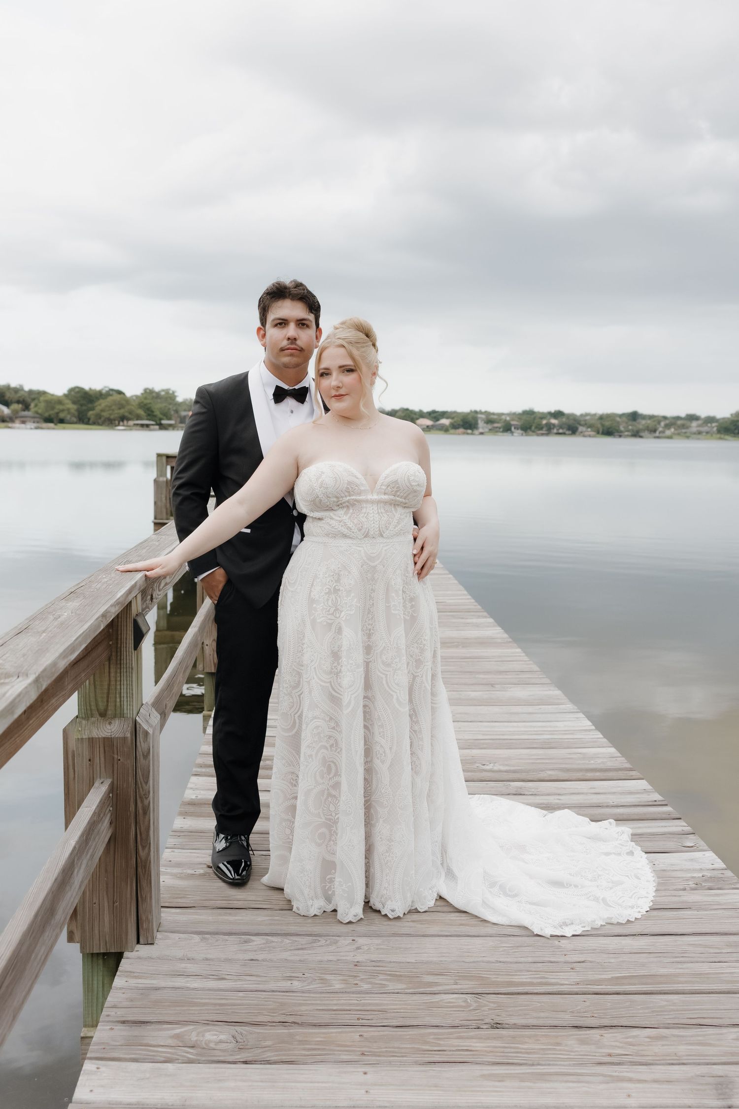 Bride and groom posing on a wooden dock by a lake, bride in lace gown, groom in black tuxedo.