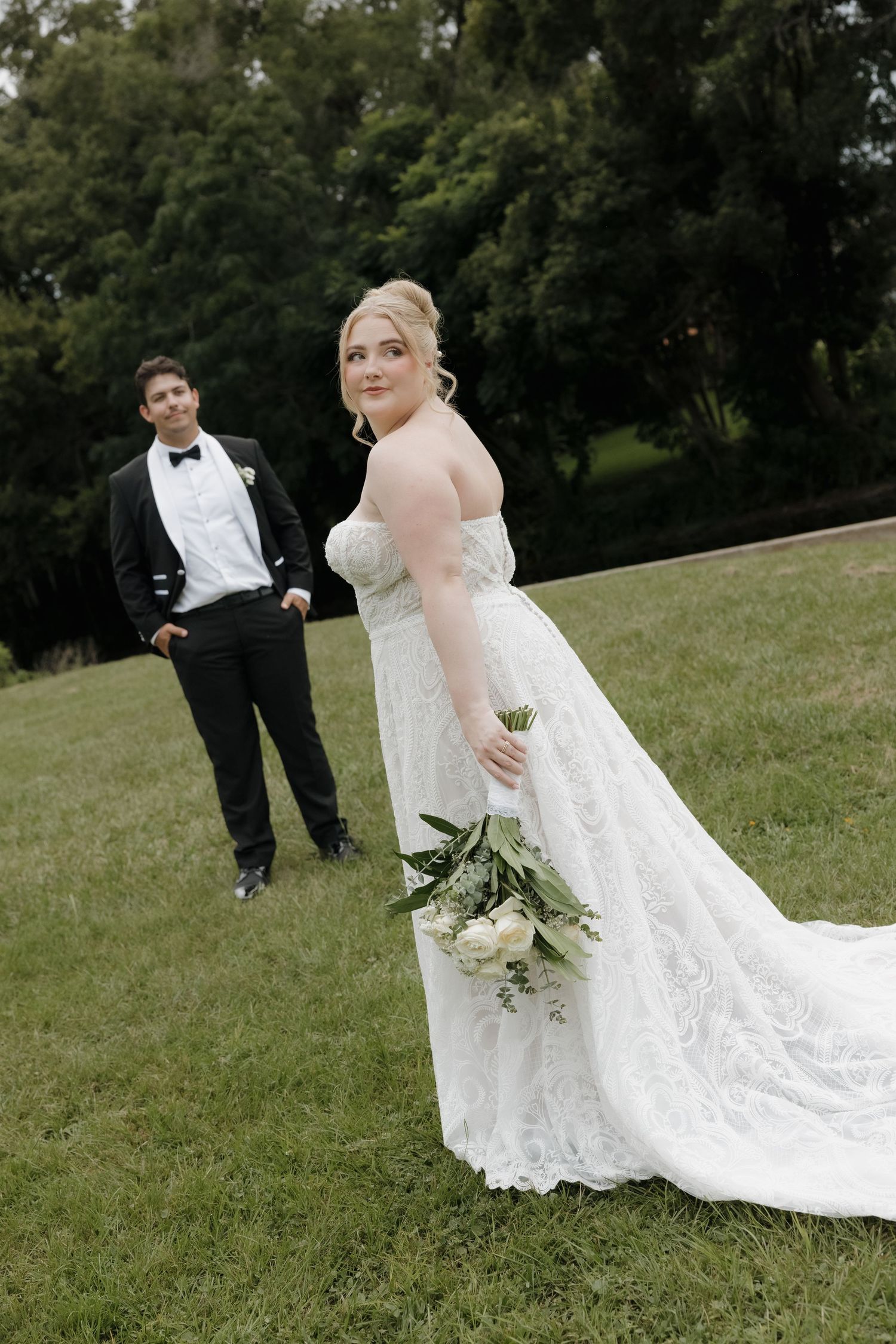 Bride in lace gown with train poses on grass while groom in tuxedo stands behind her.