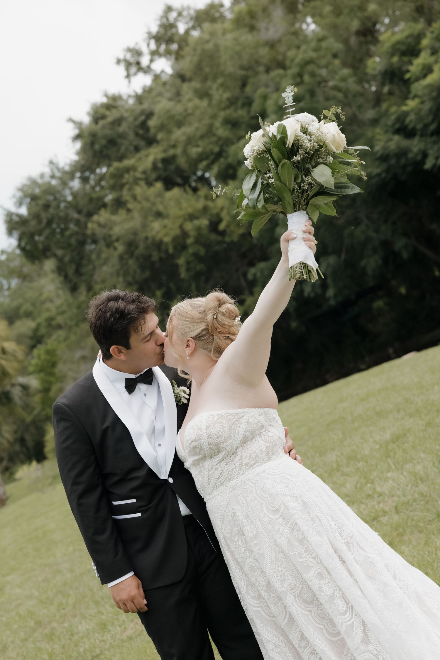 Newlyweds share a kiss outdoors as bride joyfully raises her white and green bouquet in the air.