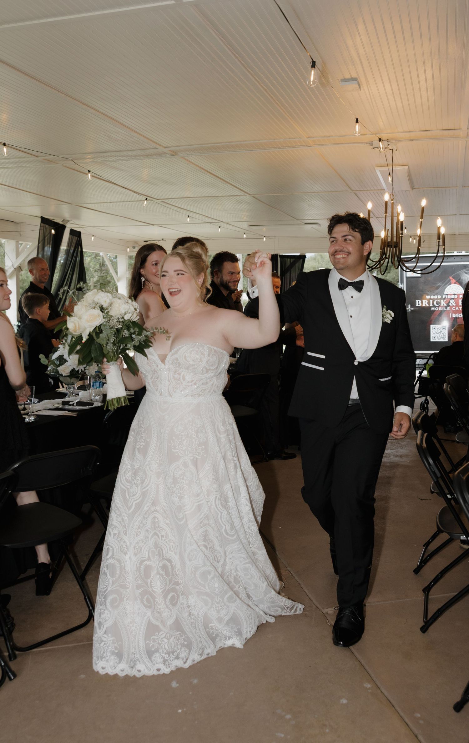Bride in lace gown and groom in tuxedo walk joyfully into wedding reception holding bouquet.
